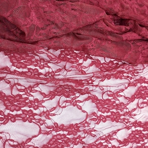 Close-up of burgundy lace fabric. The fabric is intricately patterned with floral designs and has a delicate, sheer appearance. The lace is draped, showing the texture and depth of the material. The color is a deep, rich red.