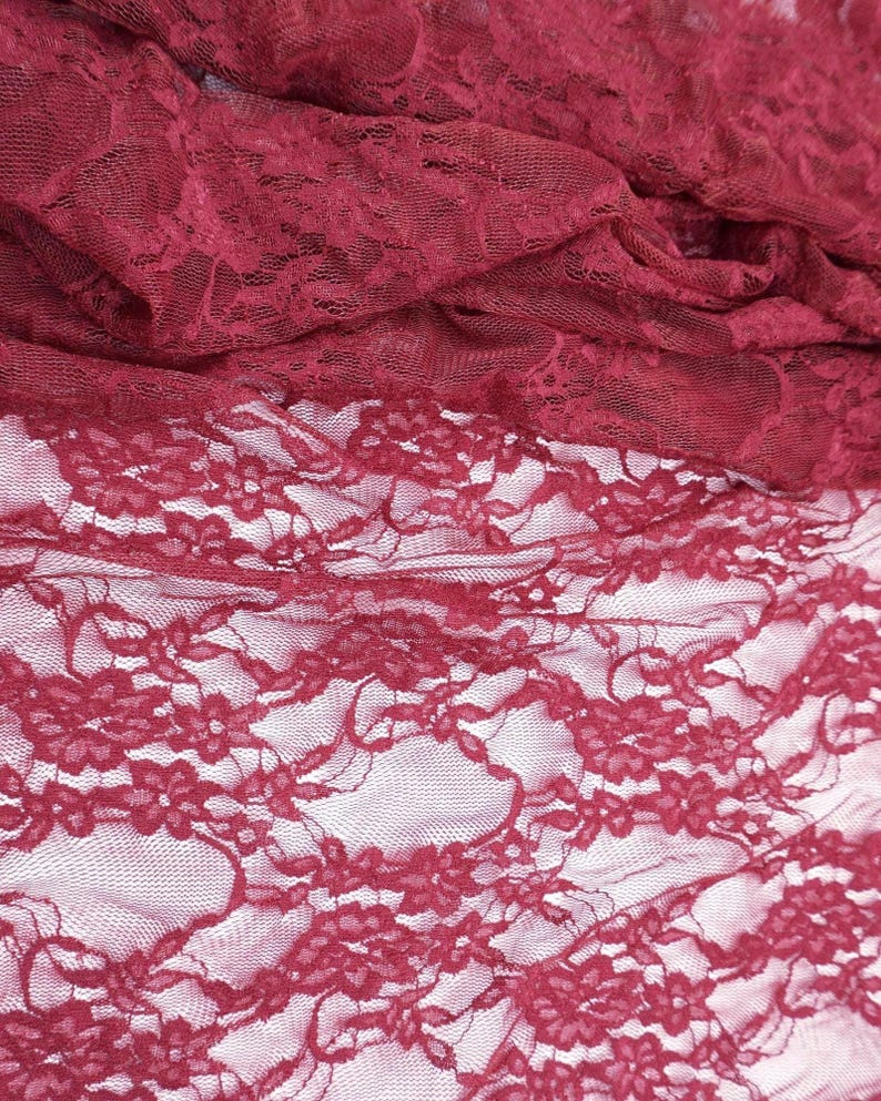 Close-up of burgundy lace fabric. The fabric is intricately patterned with floral designs and has a delicate, sheer appearance. The lace is draped, showing the texture and depth of the material. The color is a deep, rich red.