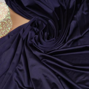 May include: A deep navy blue fabric with a smooth, matte finish draped in soft, flowing folds. To the side, a small, cylindrical spool covered in iridescent, light-reflecting sequins sits partially visible against the dark textile surface.