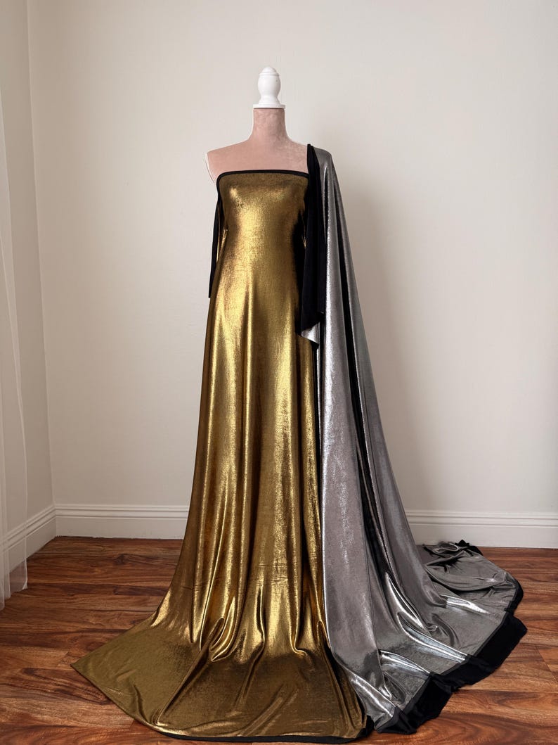 May include: A floor-length strapless gown featuring a metallic gold fabric bodice and skirt, accented by a long, flowing metallic silver train attached to the side. The garment includes black trim along the neckline and the edges of the silver fabric.