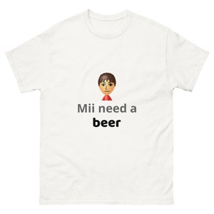 May include: White t-shirt featuring a cartoon Mii character and the phrase "Mii need a beer" in gray and black text. The Mii character has brown hair and a red collar. The shirt is a casual wear item.