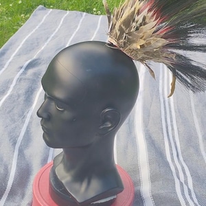 Puede incluir: Un busto de maniquí negro sobre una base roja, adornado con un tocado de plumas. El tocado presenta plumas negras, marrones y rojas. La cabeza se encuentra sobre una tela a rayas, creando un contraste visual.