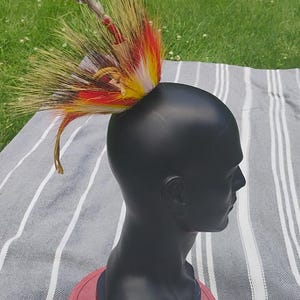 Puede incluir: Un busto de maniquí negro sobre una base roja, adornado con un tocado de plumas de colores. El tocado presenta plumas amarillas, rojas y marrones. El fondo incluye una tela a rayas y hierba verde.