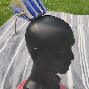 May include: A black mannequin head with a decorative headdress. The headdress has blue, white, and yellow strands. The head is on a red base, against a striped fabric and a grassy background.