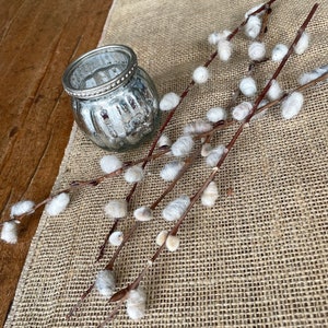 May include: A silver candle holder with a textured surface sits on a burlap table runner with several sprigs of pussy willow branches with white fluffy buds.