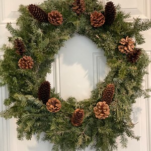May include: A green pine wreath decorated with brown pine cones. The wreath is hanging on a white door.