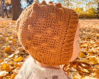 Peut inclure: Un bonnet bébé en crochet marron avec un motif texturé. Le bonnet a une longue ficelle qui peut être nouée sous le menton. Le bonnet est porté par un bébé assis sur un lit de feuilles d'automne tombées.