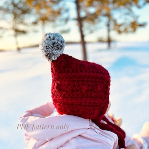 Peut inclure: Un bonnet en crochet rouge avec un pompon blanc et gris. Le bonnet est porté par une personne vêtue d'une veste rose. Le texte "PDF pattern only" est visible sur l'image.