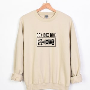 Puede incluir: Una sudadera beige con un gráfico negro de un coche de carreras y el texto "BOX BOX BOX" impreso en la parte delantera.