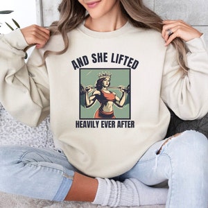May include: A cream-colored sweatshirt with the text "And She Lifted Heavily Ever After" and a graphic of a woman wearing a crown and lifting weights.
