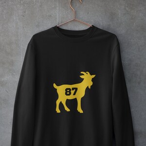 K&ouml;nnte beinhalten: Schwarzer Pullover mit einer goldenen Ziegen-Silhouette und der Nummer 87, die auf die Seite der Ziege gedruckt ist.