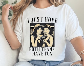 Puede incluir: Camiseta blanca con un gráfico negro de dos mujeres con sombreros sosteniendo cócteles. El texto en la camiseta dice "Solo espero que ambos equipos se diviertan."