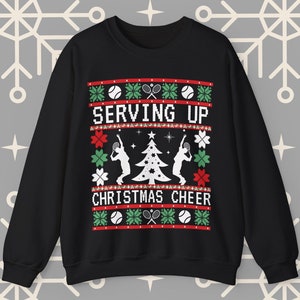 Op de afbeelding: Zwarte kersttrui met een witte kerstboom en twee tennisspelers die een tennisbal serveren. De trui heeft een rood-groen patroon met de tekst "SERVING UP CHRISTMAS CHEER".