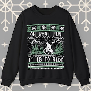 May include: Black Christmas sweater with a green and white design featuring a mountain scene with a person riding a bicycle. The text "Oh what fun it is to ride" is printed on the sweater.