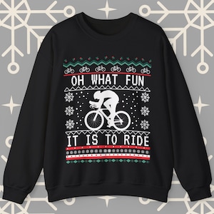 以下が含まれることがあります： 自転車に乗ったサイクリストの白いシルエットが描かれた黒いクリスマスセーター。セーターは赤と緑のパターンで、「Oh what fun it is to ride」という文字が書かれています。