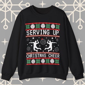 Op de afbeelding: Zwarte kersttrui met een witte volleybal en spelers, groene en rode sneeuwvlokken en de tekst "Serving Up Christmas Cheer".