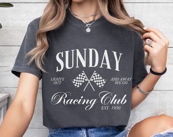 May include: A charcoal grey t-shirt with a white graphic design that says "SUNDAY LIGHTS OUT AND AWAY WE GO Racing Club EST. 1950". The graphic also includes two checkered flags crossed over each other.