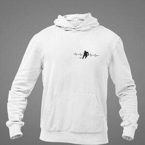 Peut inclure: Sweat-shirt à capuche blanc avec une silhouette noire d'un joueur de hockey avec un bâton de hockey et une ligne de battement de cœur.