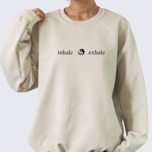 Puede incluir: Una sudadera beige con un símbolo yin yang negro y el texto "inhale exhale" impreso en la parte delantera.