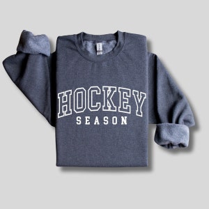 Pu&ograve; includere: Una felpa grigia scura con il testo "HOCKEY SEASON" in lettere bianche con contorno.