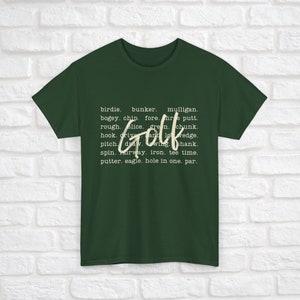 K&ouml;nnte beinhalten: Ein dunkelgr&uuml;nes T-Shirt mit dem Wort "Golf" in gro&szlig;er wei&szlig;er Schrift. Das Wort ist umgeben von kleinerem wei&szlig;em Text, der verschiedene Golfbegriffe auflistet, wie "Birdie", "Bunker", "Mulligan", "Bogey", "Chip", "Fore", "Putt", "Rough", "Slice", "Green", "Chunk", "Hook", "Drive", "Wedge", "Pitch", "Draw", "Wing", "Shank", "Spin", "Fairway", "Eisen", "Tee Time", "Putter", "Eagle", "Hole in One" und "Par".