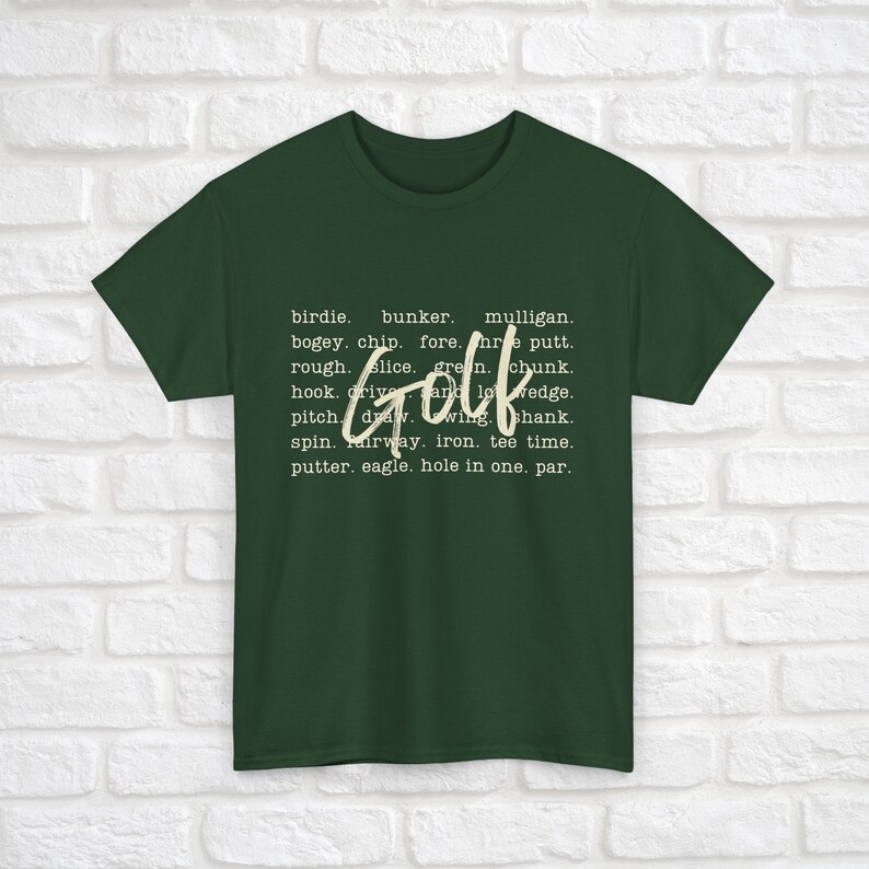 K&ouml;nnte beinhalten: Ein dunkelgr&uuml;nes T-Shirt mit dem Wort "Golf" in gro&szlig;er wei&szlig;er Schrift. Das Wort ist umgeben von kleinerem wei&szlig;em Text, der verschiedene Golfbegriffe auflistet, wie "Birdie", "Bunker", "Mulligan", "Bogey", "Chip", "Fore", "Putt", "Rough", "Slice", "Green", "Chunk", "Hook", "Drive", "Wedge", "Pitch", "Draw", "Wing", "Shank", "Spin", "Fairway", "Eisen", "Tee Time", "Putter", "Eagle", "Hole in One" und "Par".