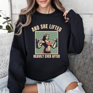 May include: Black sweatshirt with a vintage-style graphic of a woman wearing a crown and lifting weights. The text reads "And She Lifted Heavily Ever After."