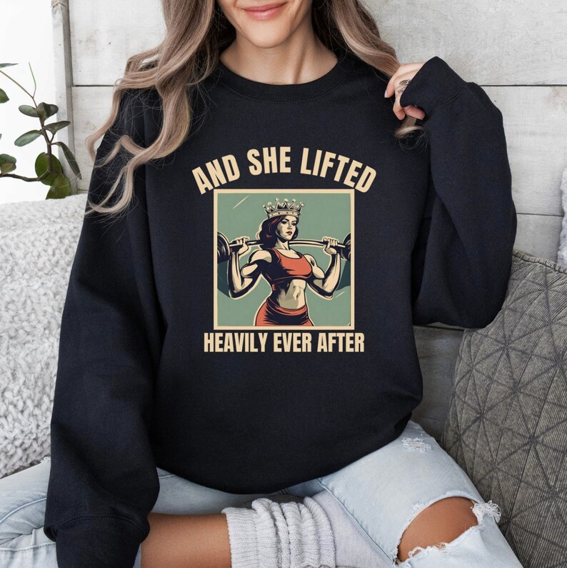 May include: Black sweatshirt with a vintage-style graphic of a woman wearing a crown and lifting weights. The text reads "And She Lifted Heavily Ever After."
