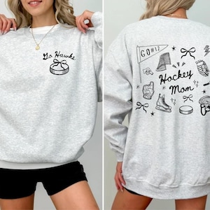 Woman wearing a crewneck sweatshirt with “Hockey Mom” doodle graphic on the back and personalized team name "Go Hawks" and player number "12" on the front; casual hockey game day outfit for moms.