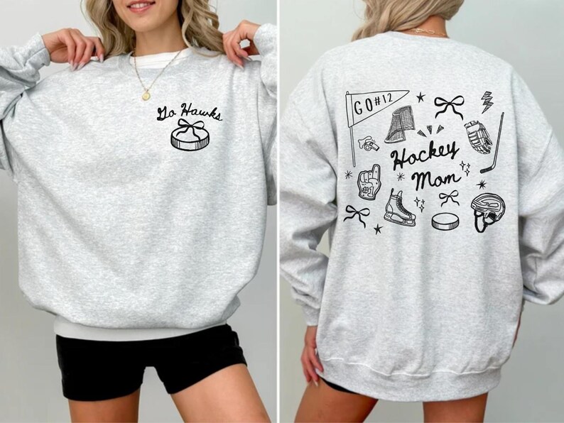 Woman wearing a crewneck sweatshirt with “Hockey Mom” doodle graphic on the back and personalized team name "Go Hawks" and player number "12" on the front; casual hockey game day outfit for moms.