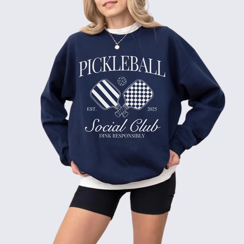 Puede incluir: Una sudadera azul marino con un dise&ntilde;o gr&aacute;fico blanco que dice "Pickleball Social Club Est. 2025 Dink Responsibly" con dos palas de pickleball cruzadas.