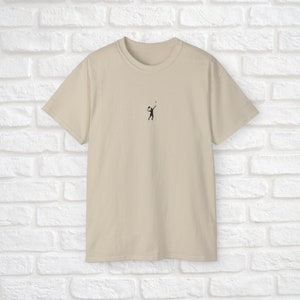Pu&ograve; includere: Una t-shirt beige con una piccola silhouette nera di una persona che gioca a pallavolo sul davanti.