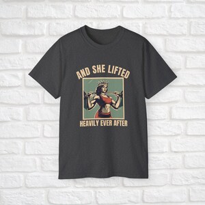 Może przedstawiać: Szary t-shirt z grafiką w stylu vintage przedstawiającą kobietę w koronie podnoszącą sztangę. Pod grafiką znajduje się napis "And She Lifted Heavily Ever After".