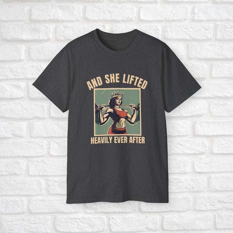 Może przedstawiać: Szary t-shirt z grafiką w stylu vintage przedstawiającą kobietę w koronie podnoszącą sztangę. Pod grafiką znajduje się napis "And She Lifted Heavily Ever After".