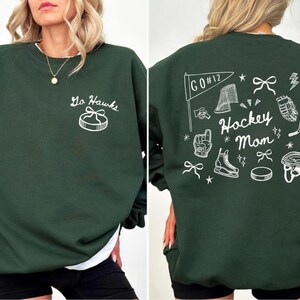 Woman wearing a crewneck sweatshirt with “Hockey Mom” doodle graphic on the back and personalized team name "Go Hawks" and player number "12" on the front; casual hockey game day outfit for moms.