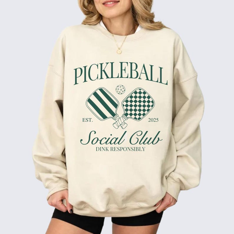 Puede incluir: Una sudadera beige con un dise&ntilde;o gr&aacute;fico verde que dice "Pickleball Social Club Est. 2025 Dink Responsibly". El gr&aacute;fico incluye dos palas de pickleball y una pelota.