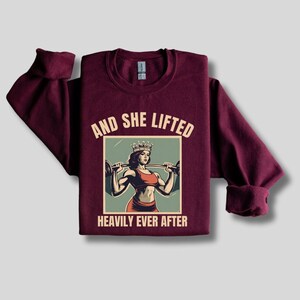 May include: A maroon sweatshirt with a vintage-style graphic of a woman wearing a crown and lifting weights. The text "And She Lifted Heavily Ever After" is printed below the graphic.