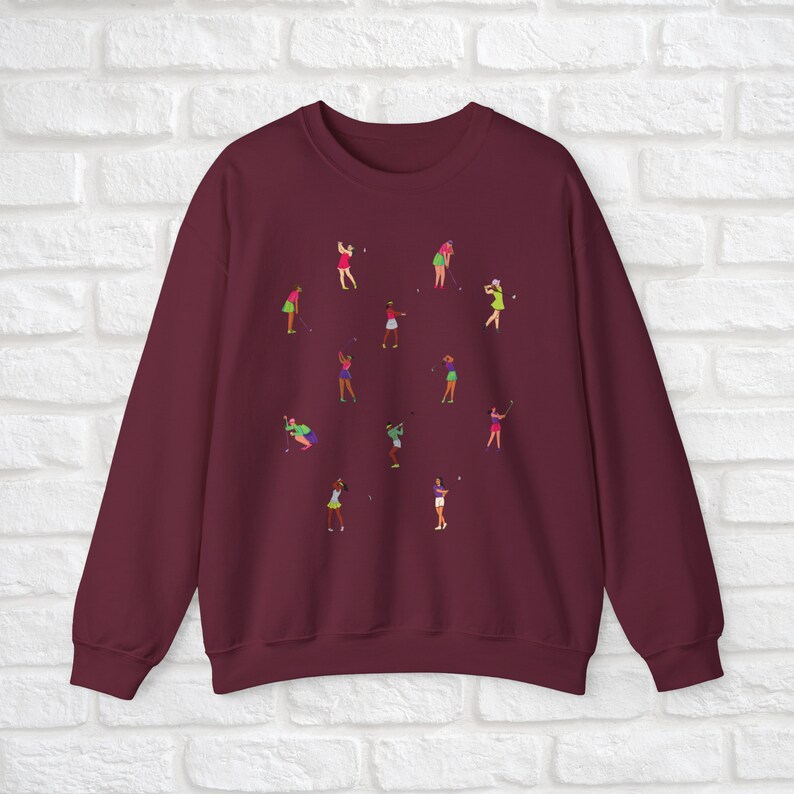 May include: Burgundy sweatshirt featuring a design of women playing golf. The women are depicted in various poses and wearing different coloured clothing.