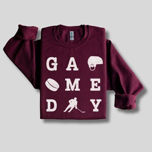 K&ouml;nnte beinhalten: Ein weinroter Pullover mit den Worten "GAME DAY" in wei&szlig;en Buchstaben. Die Buchstaben sind von Hockey-Symbolen umgeben, darunter ein Puck, ein Schl&auml;ger und ein Helm.