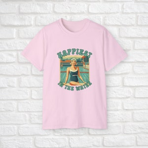 May include: Pink t-shirt with a retro graphic of a woman in a swimming pool. The graphic says "Happiest in the water".