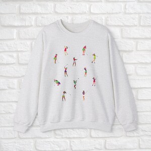 May include: A grey sweatshirt featuring a colourful design of women playing golf. The design depicts multiple women in various poses, swinging golf clubs and holding golf balls.