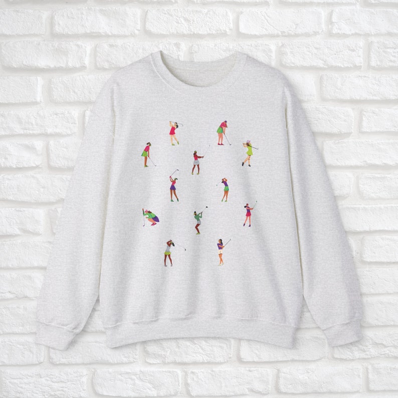 May include: A grey sweatshirt featuring a colourful design of women playing golf. The design depicts multiple women in various poses, swinging golf clubs and holding golf balls.