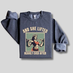 May include: A dark blue sweatshirt with the text "And She Lifted Heavily Ever After" and a vintage-style illustration of a woman wearing a crown and lifting a barbell.