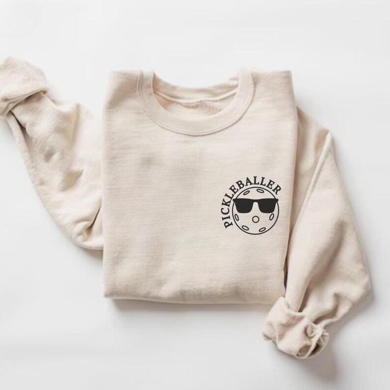 May include: A beige sweatshirt with a black graphic of a pickleball with sunglasses and the text "Pickleballer" on the front.