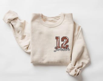 May include: A cream-coloured sweatshirt with a crew neck. The sweatshirt features a football-themed design with the number 12 and the name "Smith" in a brown and white colour scheme. The sleeves are rolled up.