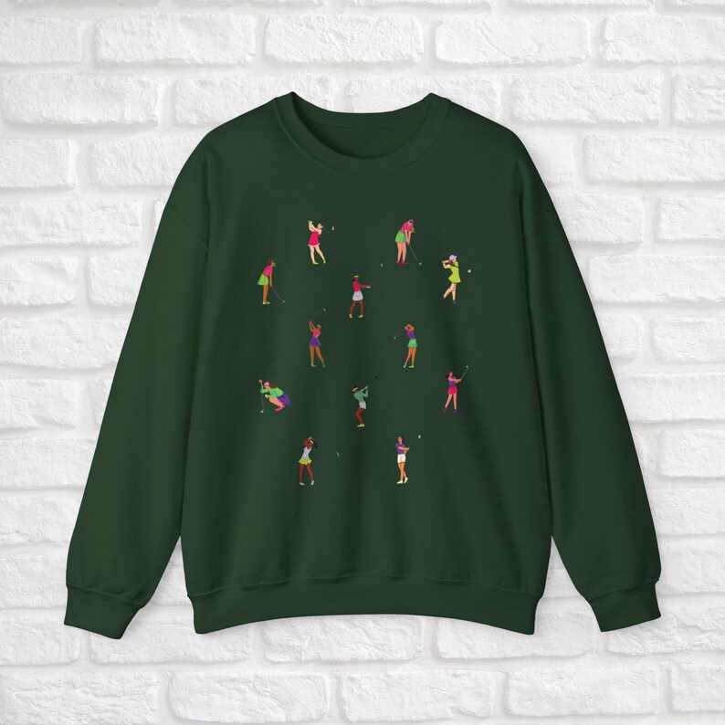 May include: A dark green sweatshirt featuring a design of women playing golf. The women are depicted in various poses and wearing different coloured clothing.
