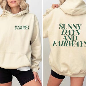 beige golf themed hoodie with sunny days and fairways in a bold, trendy font on the back of the shirt. On the front, is a small sunny days and fairways graphic on the left chest