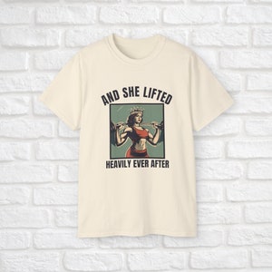 Może przedstawiać: Kremowy t-shirt z grafiką kobiety w koronie podnoszącej ciężary. Pod grafiką znajduje się napis "And she lifted heavily ever after".