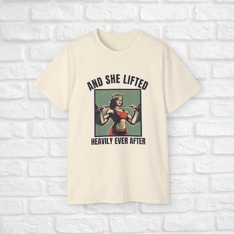 Może przedstawiać: Kremowy t-shirt z grafiką kobiety w koronie podnoszącej ciężary. Pod grafiką znajduje się napis "And she lifted heavily ever after".