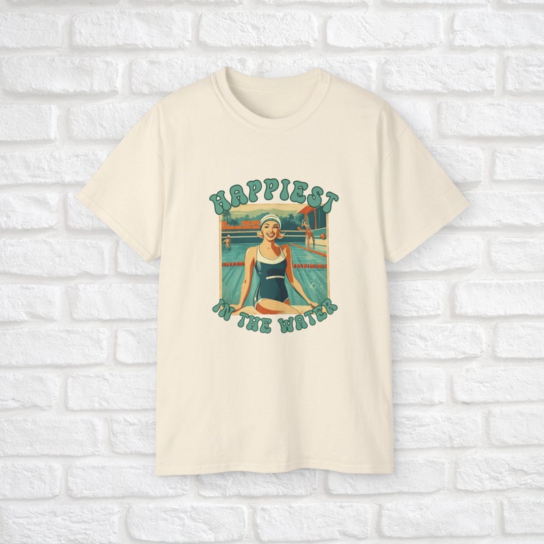 May include: A cream colored t-shirt with a retro graphic design featuring a woman in a blue swimsuit sitting by a pool. The text on the graphic reads "Happiest in the Water".